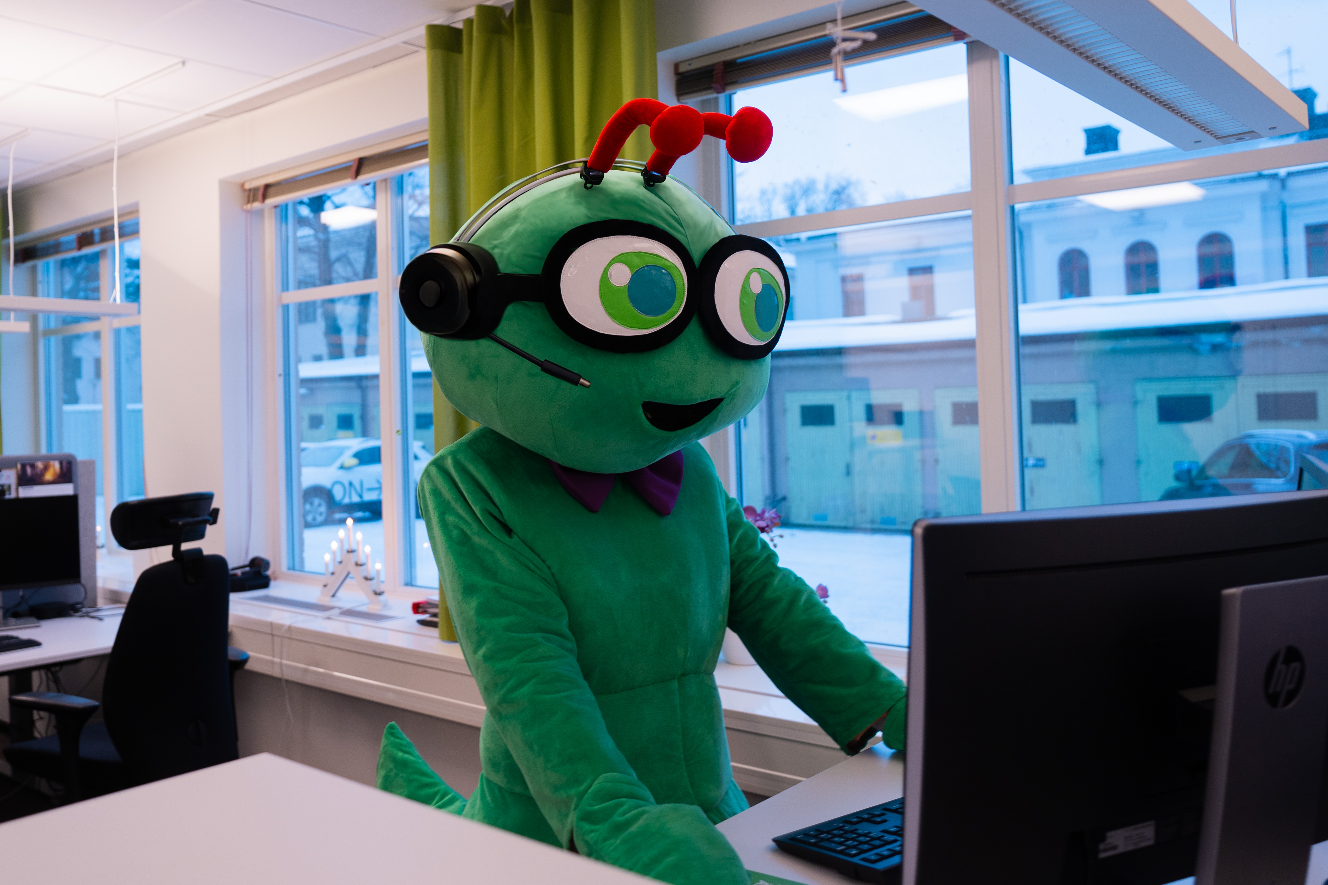 Sörmlandstrafikens gröna maskot Busse står bakom en dator med headset och pratar i telefon.
